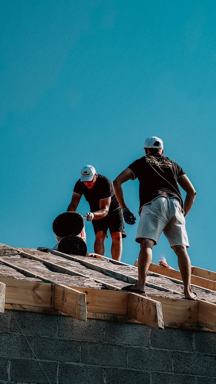 about-us Two construction workers apply roofing materials under a bright blue sky, showcasing teamwork and labor.